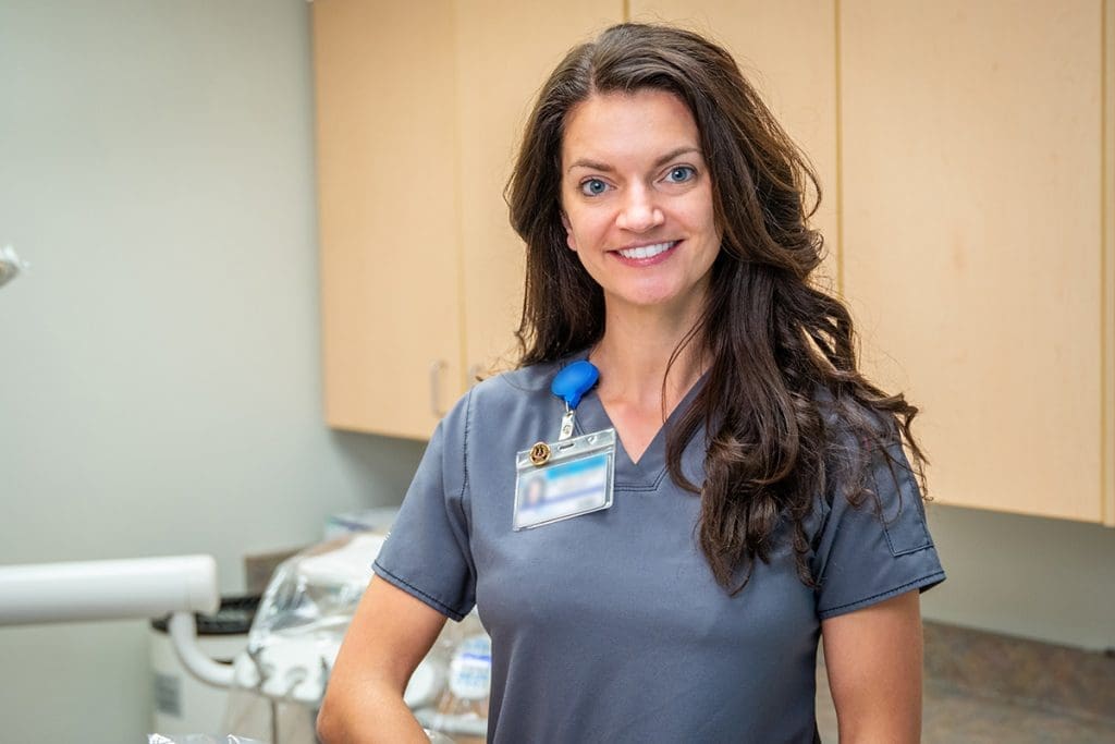 A woman in gray medical scrubs smiles at the camera in a clinic room, wearing a badge with a blue clip, representing Grand Junction Health Care, with cabinets and medical equipment in the background.