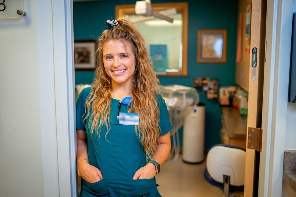 A smiling woman in teal medical scrubs stands in a doorway at Grand Junction Health Care, hands in her pockets. Her long wavy hair and ID badge are visible, with a brightly lit clinic and medical equipment behind her.