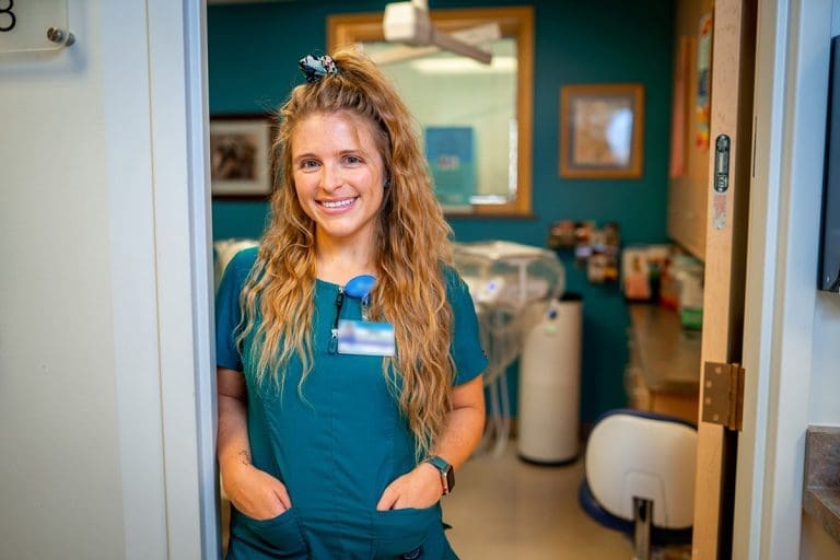 A smiling woman in teal medical scrubs stands in a doorway at Grand Junction Health Care, hands in her pockets. Her long wavy hair and ID badge are visible, with a brightly lit clinic and medical equipment behind her.