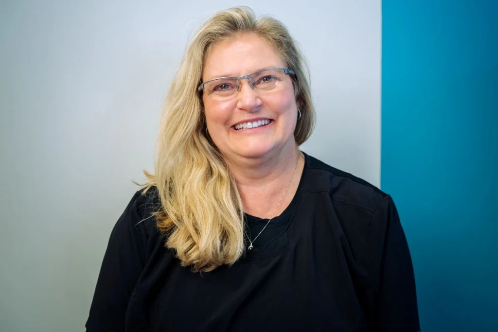 A smiling woman with long blonde hair and glasses, wearing a black top, stands in front of a wall with white and teal blue sections, reflecting the welcoming atmosphere often found in Grand Junction Health Care settings.