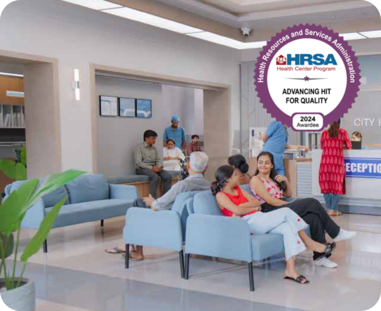 People sit and talk in a bright Grand Junction Health Care clinic waiting area with blue chairs and a reception desk; a large HRSA 2024 Advancing HIT For Quality award badge is displayed in the top right corner.