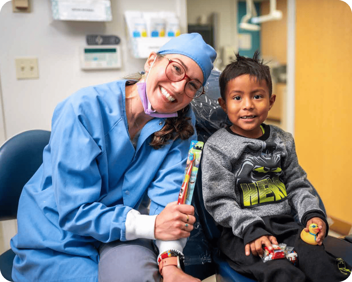 A smiling dental professional in blue scrubs and a surgical cap sits beside a young boy in a dental chair. Both are smiling; the dental professional holds a toothbrush, showing their commitment to affordable health care. The boy holds a toy.