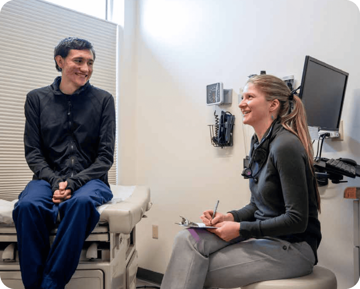 A patient sits on an exam table smiling while a Montrose Health Care provider sits nearby, holding a clipboard and pen, also smiling. Medical equipment and a computer are visible in the exam room, reflecting quality, affordable health care.
