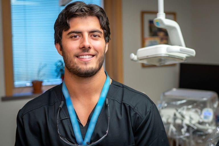 A young male dentist in black scrubs smiles in a Montrose Health Care dental office, with dental equipment and an examination chair visible in the background.