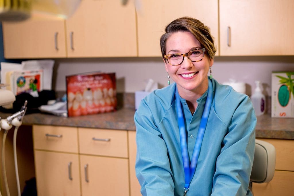 A smiling woman with short hair and glasses, wearing a teal dental coat and blue lanyard, sits in a dental office with wooden cabinets and dental supplies—reflecting the friendly atmosphere of Grand Junction Health Care.