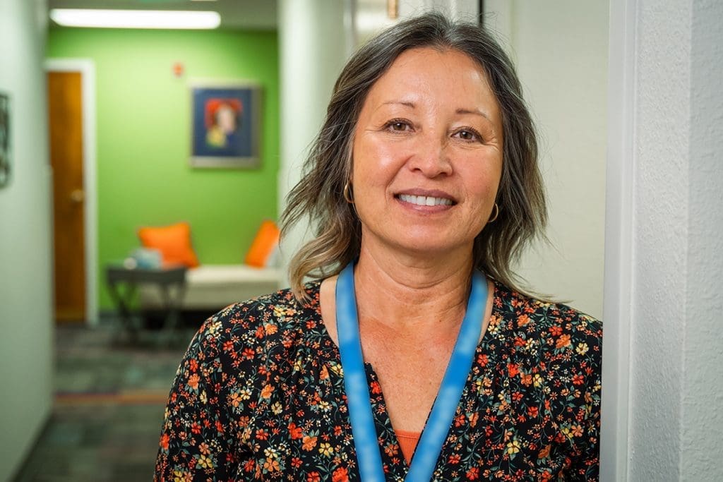 A woman with shoulder-length hair and a floral top smiles while standing in a hallway at Montrose Health Care. The background features green walls, a bench with orange pillows, and framed artwork.