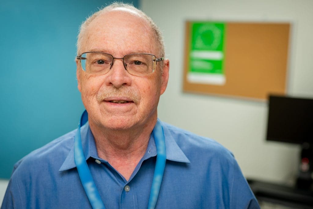 An older man with glasses, wearing a blue collared shirt and a Montrose Health Care lanyard, stands in an office with a blurred bulletin board and computer monitor in the background.