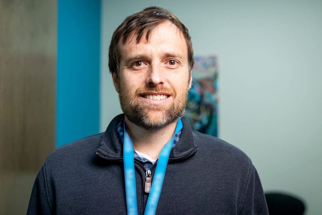 A man with brown hair and a beard, smiling, wearing a dark zip-up jacket and a teal lanyard, stands indoors against a light blue wall with blurred artwork in the background at Montrose Health Care.