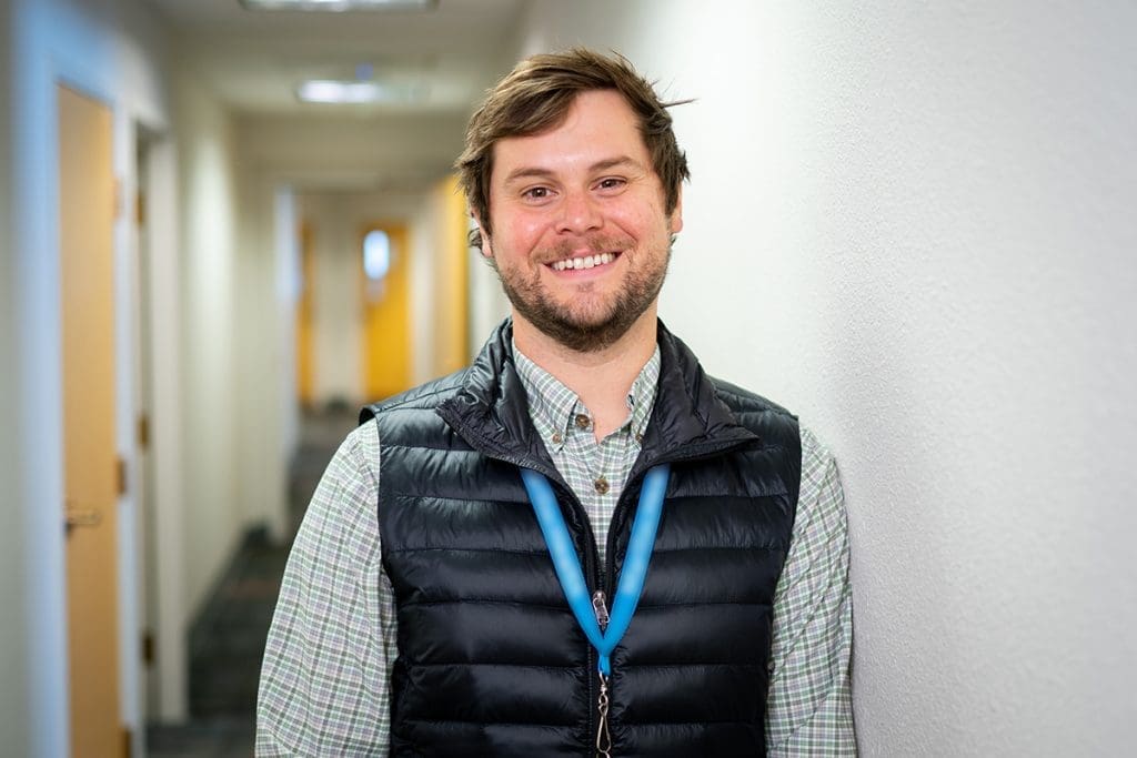 A man with short brown hair and a beard, wearing a plaid shirt, dark vest, and a blue lanyard, stands smiling against a white wall in a brightly lit Grand Junction Health Care hallway.