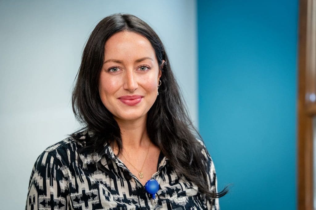 A woman with long dark hair and blue eyes, wearing a patterned black and white shirt, stands smiling in front of a blue and white background at Montrose Health Care.