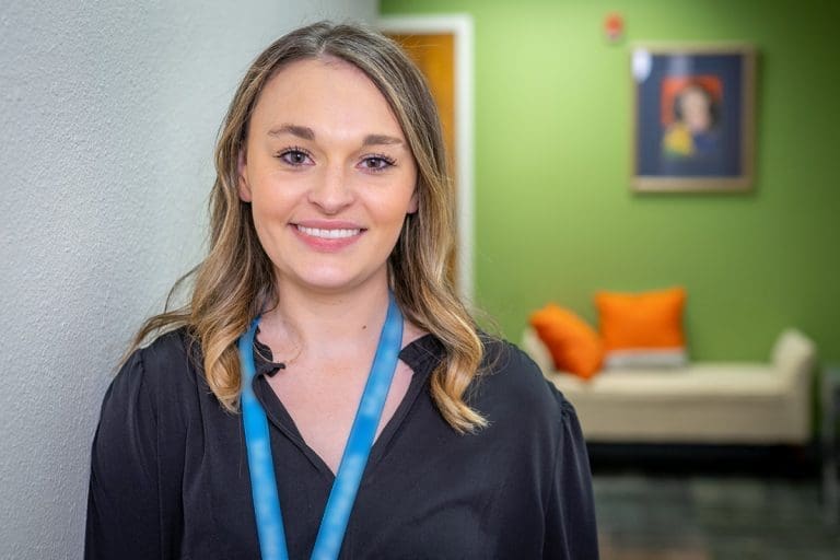 A woman with light brown hair, wearing a black top and a teal lanyard, smiles while standing indoors near a white wall. Behind her is a green wall, a couch with orange pillows, and a framed picture—reflecting Affordable Health Care values.