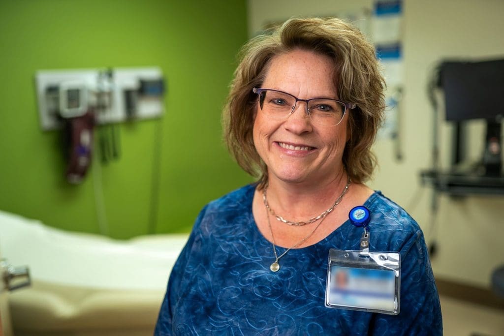 A woman wearing glasses and a blue blouse stands smiling in a Montrose Health Care office, with medical equipment on the wall behind her. She has shoulder-length hair and wears an ID badge.