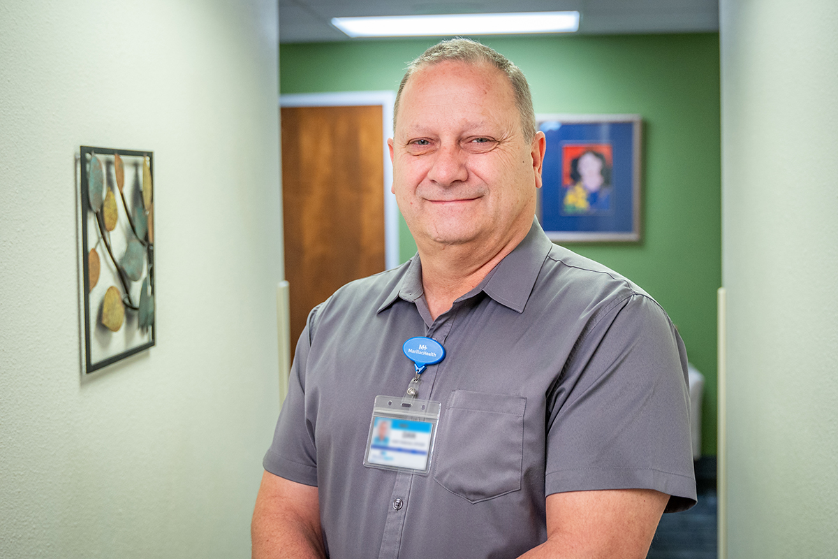 A smiling man wearing a gray short-sleeve shirt and an ID badge stands in a hallway with green walls, framed art, and a brown door in the background at Grand Junction Health Care.