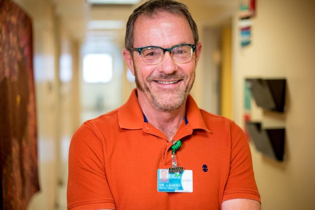 A smiling man with glasses and a beard stands in a hallway at Montrose Health Care, wearing an orange polo shirt and a visible ID badge. The softly blurred background shows doors and wall-mounted file holders, reflecting a professional healthcare setting.