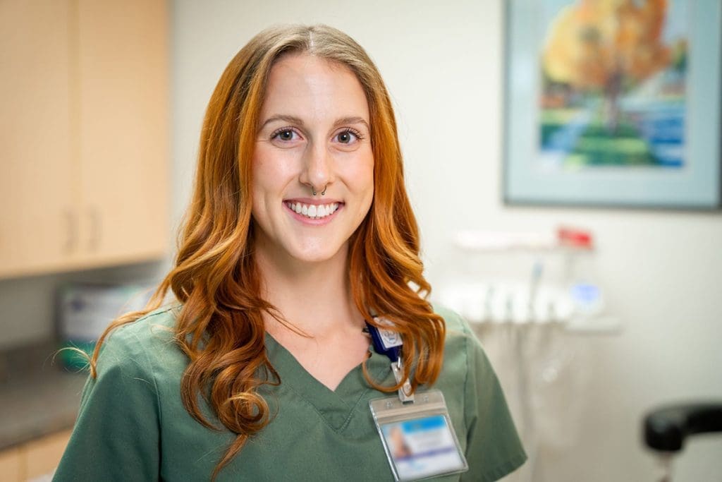 A woman with long, wavy auburn hair wearing green scrubs and a name badge smiles in a medical office, representing Grand Junction Health Care, with cabinets and a colorful painting in the background.