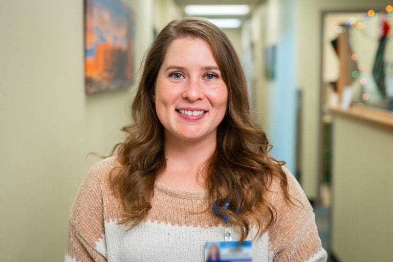 A woman with long brown hair wearing a beige and white sweater smiles at the camera in a brightly lit hallway at Montrose Health Care, with art on the walls and a decorated office area in the background.