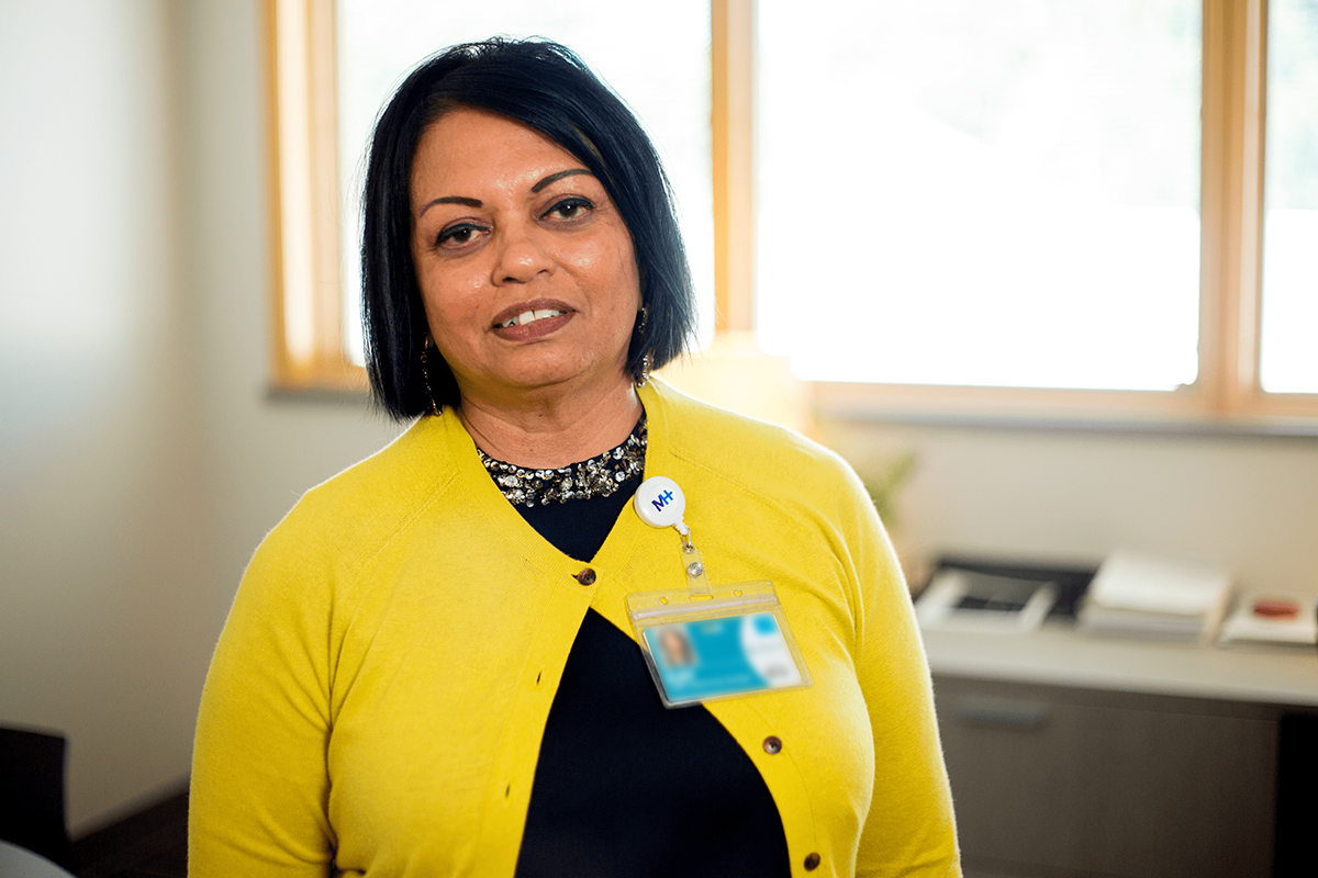A woman with shoulder-length dark hair wearing a yellow cardigan and a Montrose Health Care name badge stands in an office with large windows and a desk in the background.