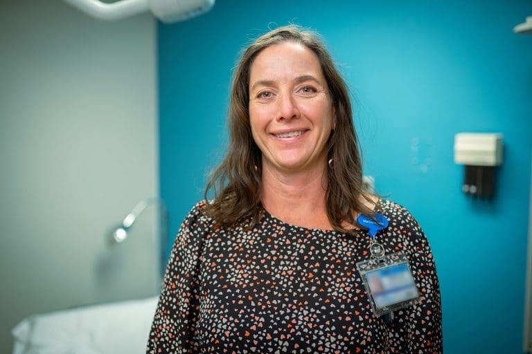 A woman with long brown hair, wearing a patterned blouse and an ID badge, stands smiling in a Montrose Health Care office with teal and gray walls and medical equipment in the background.