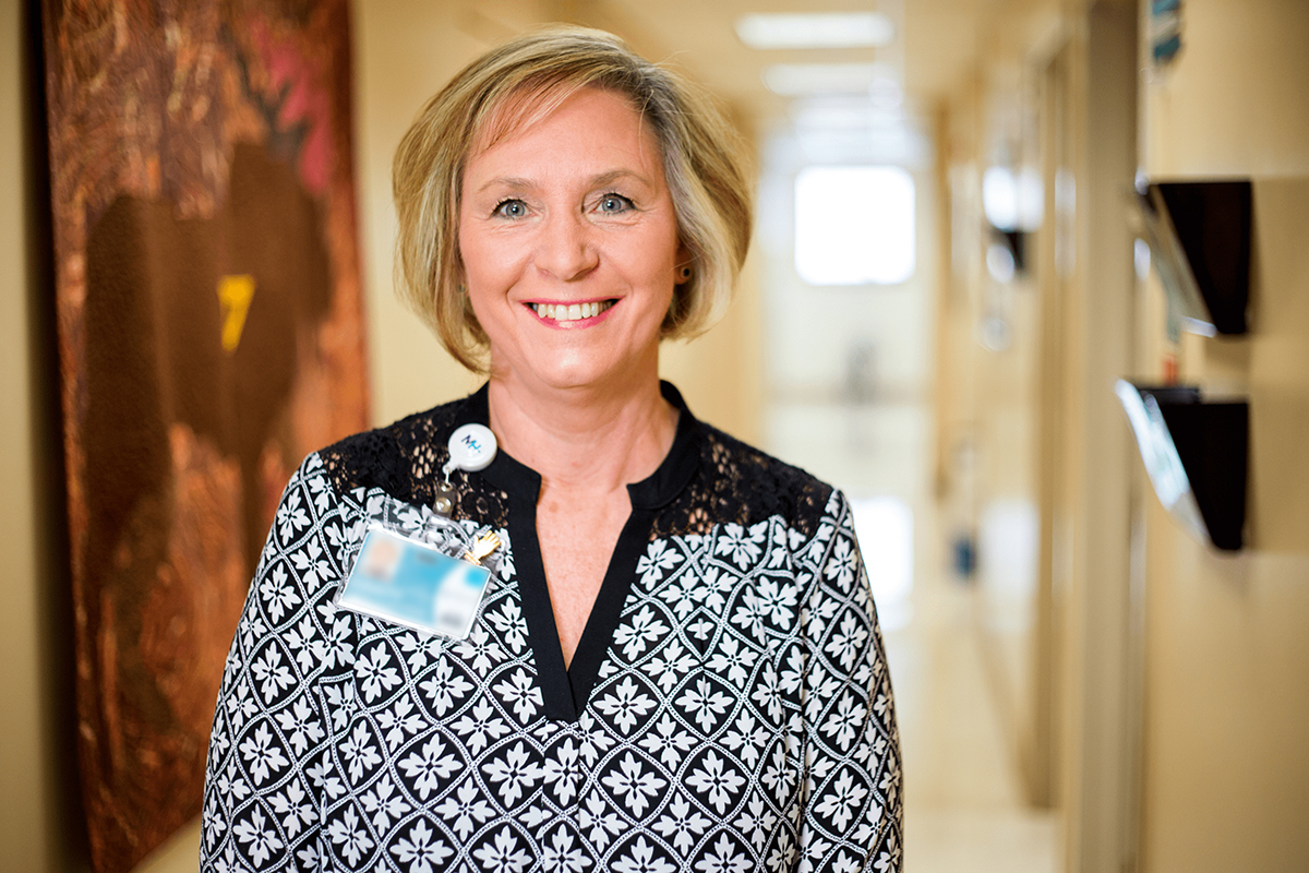 Smiling woman with short blonde hair wearing a patterned blouse and blurred ID badge stands in a brightly-lit Grand Junction Health Care hallway, with art on the wall in the background.