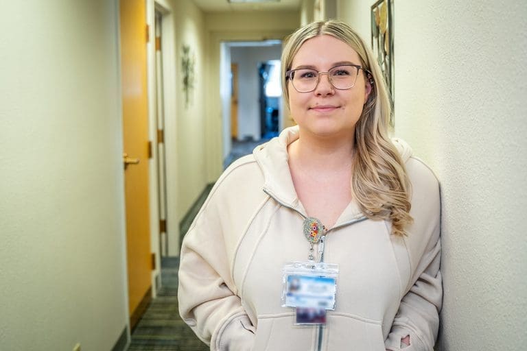 A woman with long blonde hair and glasses, wearing a light-colored hoodie and an ID badge, stands in a hallway with beige walls and office doors at a healthcare facility.