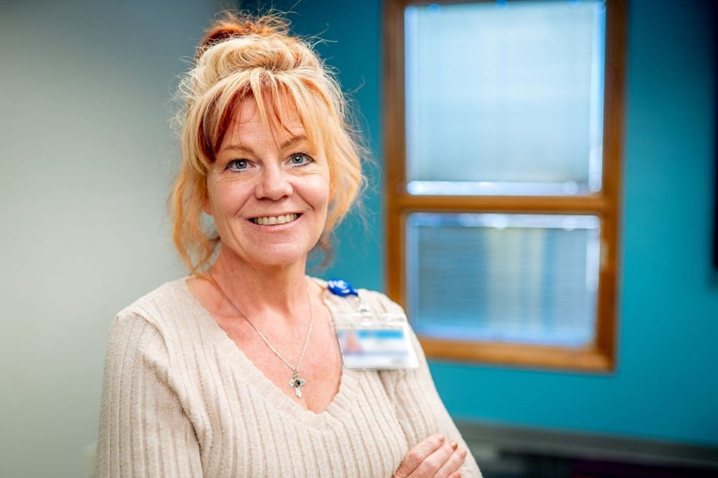 A woman with light red hair tied up, wearing a beige sweater and an ID badge, smiles while standing indoors in front of blue and white walls—reflecting her commitment to affordable health care.