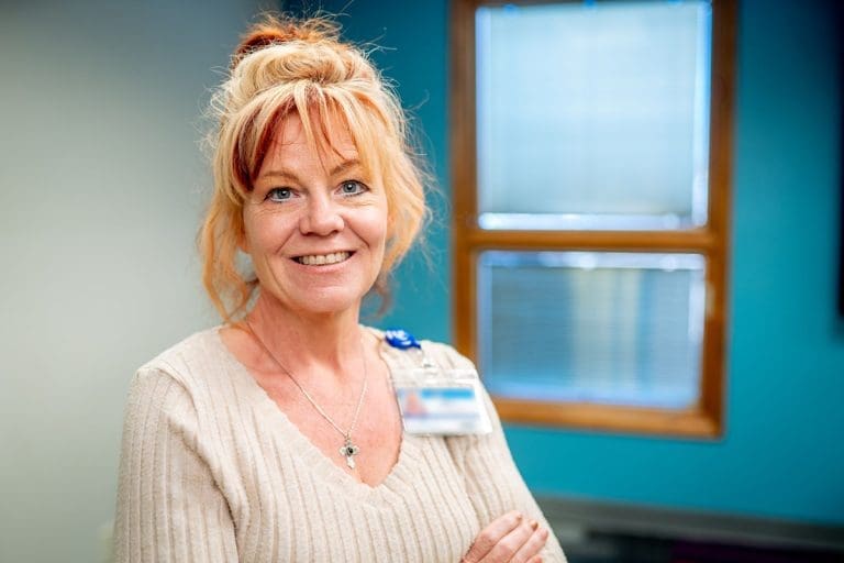 A woman with light red hair tied up, wearing a beige sweater and an ID badge, smiles while standing indoors in front of blue and white walls—reflecting her commitment to affordable health care.