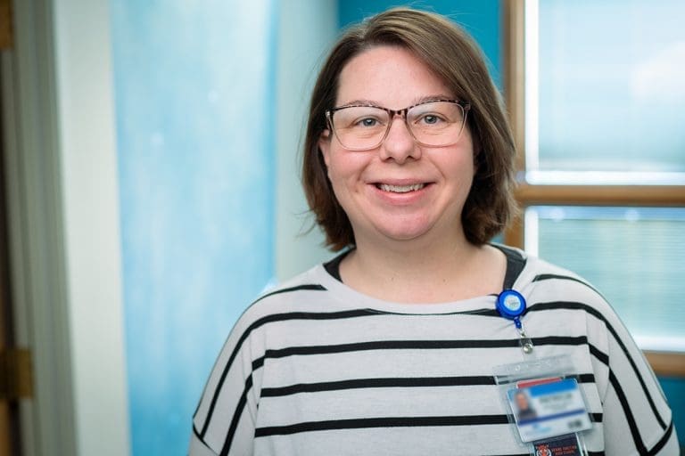 A woman with glasses and shoulder-length brown hair, wearing a white and black striped shirt and a name badge, smiles in a bright indoor setting at Grand Junction Health Care, with blue walls and windows.