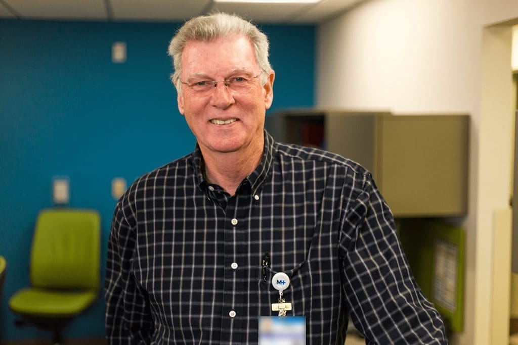 An older man wearing glasses, a black and white checkered shirt, and a work badge stands smiling in a Montrose Health Care office with blue and white walls, green chairs, and cabinets in the background.