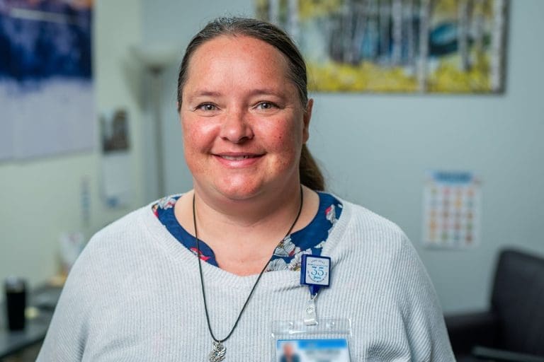 A woman with brown hair pulled back smiles at the camera. She is wearing a blue floral top and a Montrose Health Care ID badge. The background shows a blurred office with artwork, a calendar, and furniture.