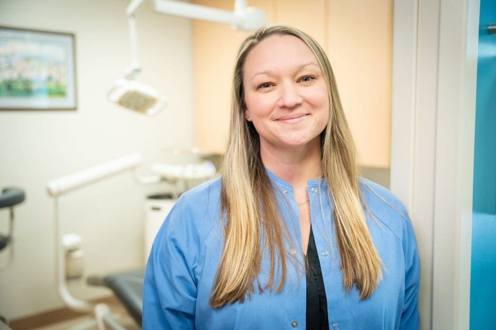 A woman with long blonde hair, wearing a light blue dental coat, smiles while standing in a dental office at Grand Junction Health Care, with dental equipment and a chair in the background.