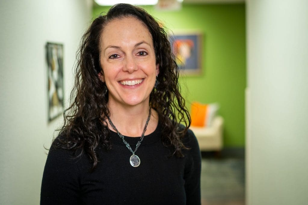 A woman with long curly dark hair, wearing a black top and a necklace, smiles at the camera in a hallway with green walls and artwork, reflecting the welcoming atmosphere of Grand Junction Health Care.
