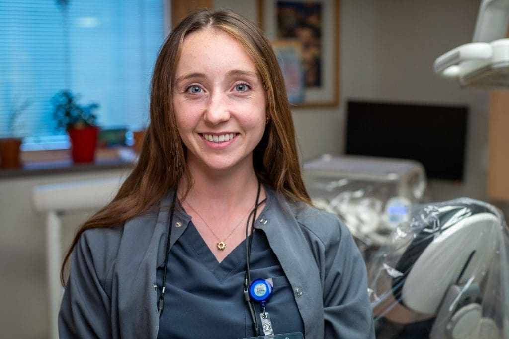 A woman in medical scrubs and a name badge smiles in a clinical office setting, surrounded by medical equipment and a window with potted plants, reflecting a welcoming environment dedicated to affordable health care.