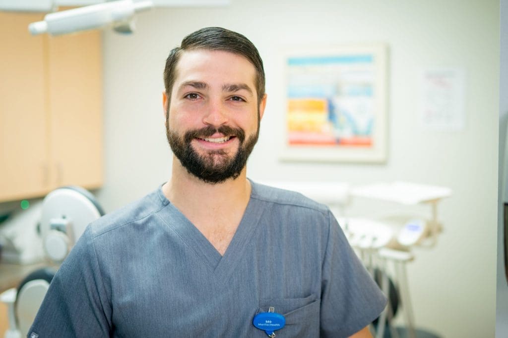 A man with a beard, wearing gray scrubs and a blue name badge, smiles while standing in a brightly lit healthcare office. Dental equipment and colorful wall art are visible in the background, reflecting a commitment to affordable health care.
