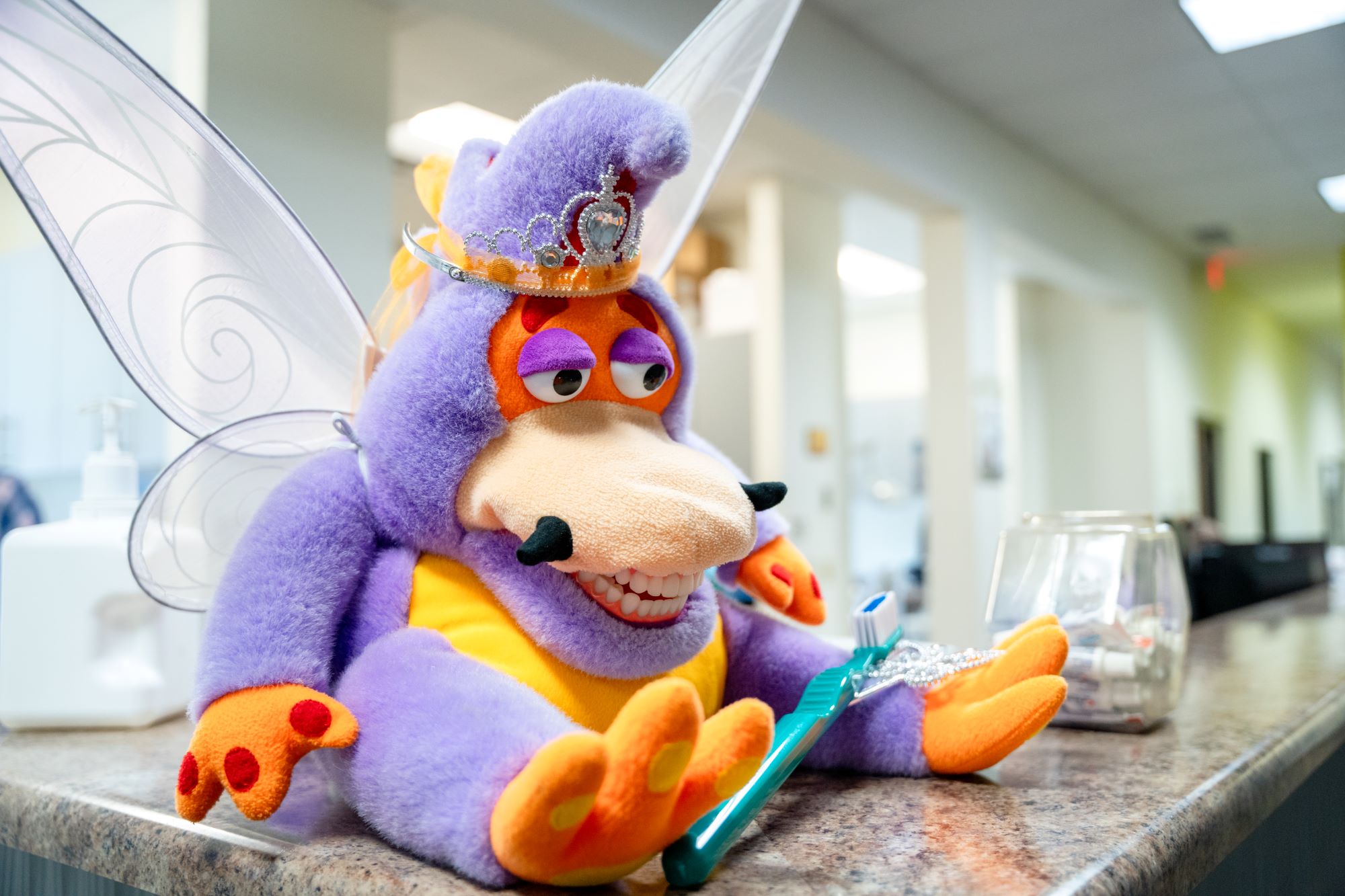 A purple and orange dragon plush toy with fairy wings and a tiara sits on a counter, holding a green toothbrush. The bright, modern office hallway suggests a playful touch in a Grand Junction Health Care or Healthcare clinic setting.