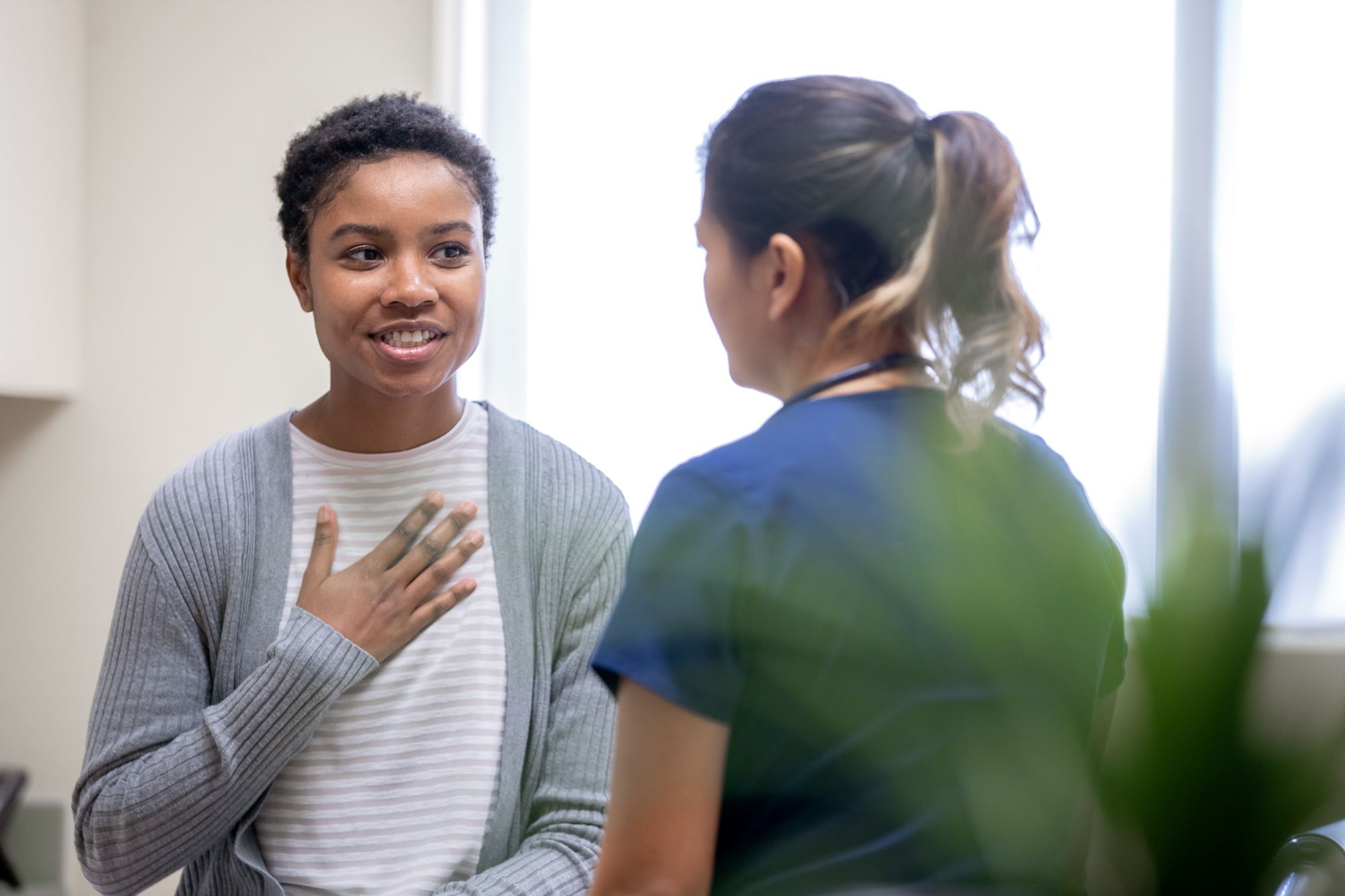 A patient sits on an exam table, smiling and touching their chest while talking with a Montrose Health Care professional in scrubs, in a well-lit medical office.