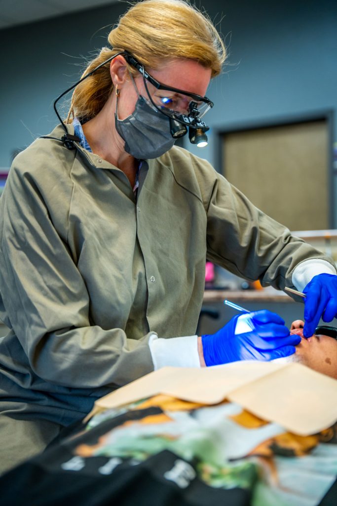 A dentist wearing magnifying glasses, a face mask, and gloves examines a patients mouth with dental tools in a Grand Junction Health Care clinic setting.