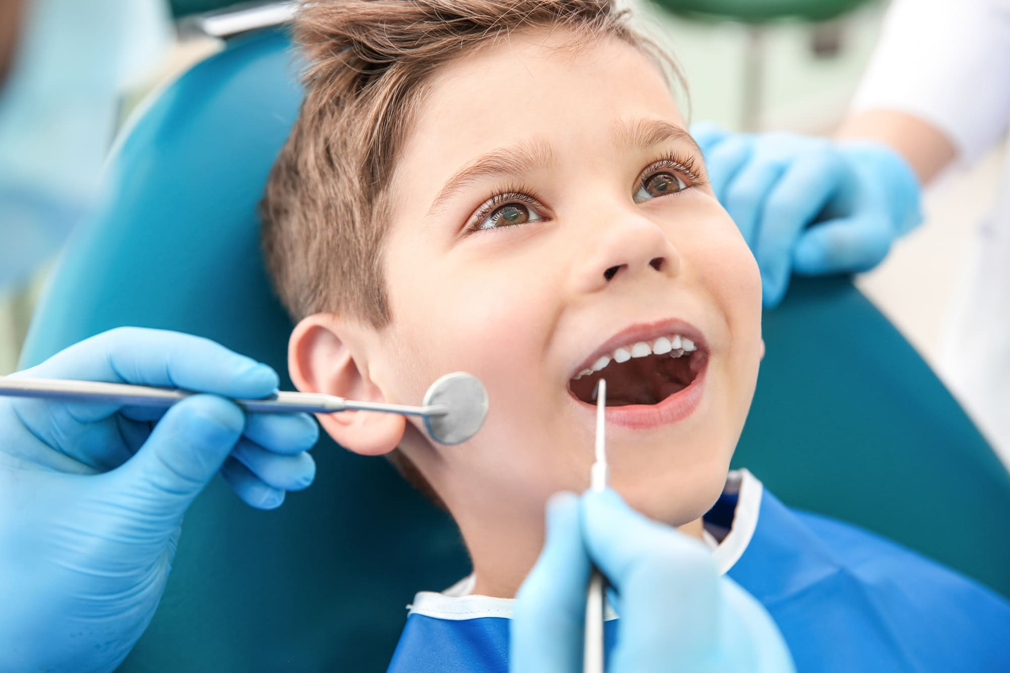A young boy sits in a dental chair, smiling with his mouth open as two gloved dentists from Grand Junction Health Care examine his teeth using dental instruments.