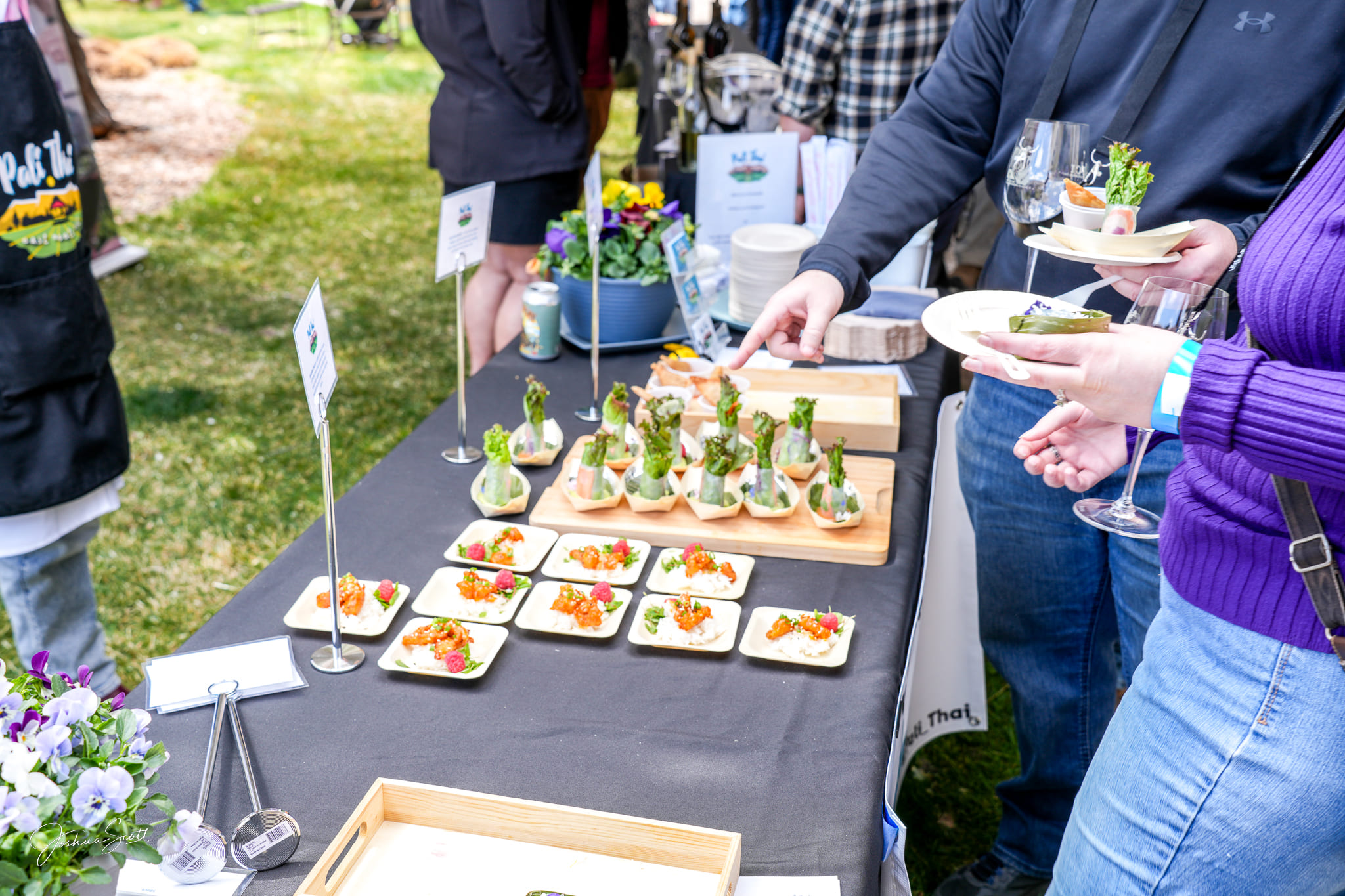 People stand at an outdoor food event, selecting appetizers from a table covered with small plates of colorful hors d’oeuvres and vibrant flower arrangements—an occasion sponsored by Montrose Health Care. One person holds a plate and a wine glass.