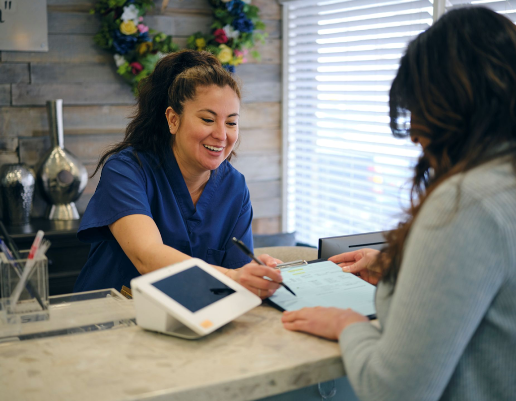A smiling receptionist in blue scrubs helps a woman fill out paperwork at the front desk of Montrose Health Care, a bright office with a floral wreath and desk accessories in the background.