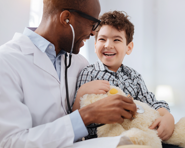 A smiling doctor uses a stethoscope on a teddy bear held by a laughing young boy, both sharing a joyful moment during a Montrose Health Care checkup in a bright, friendly office.
