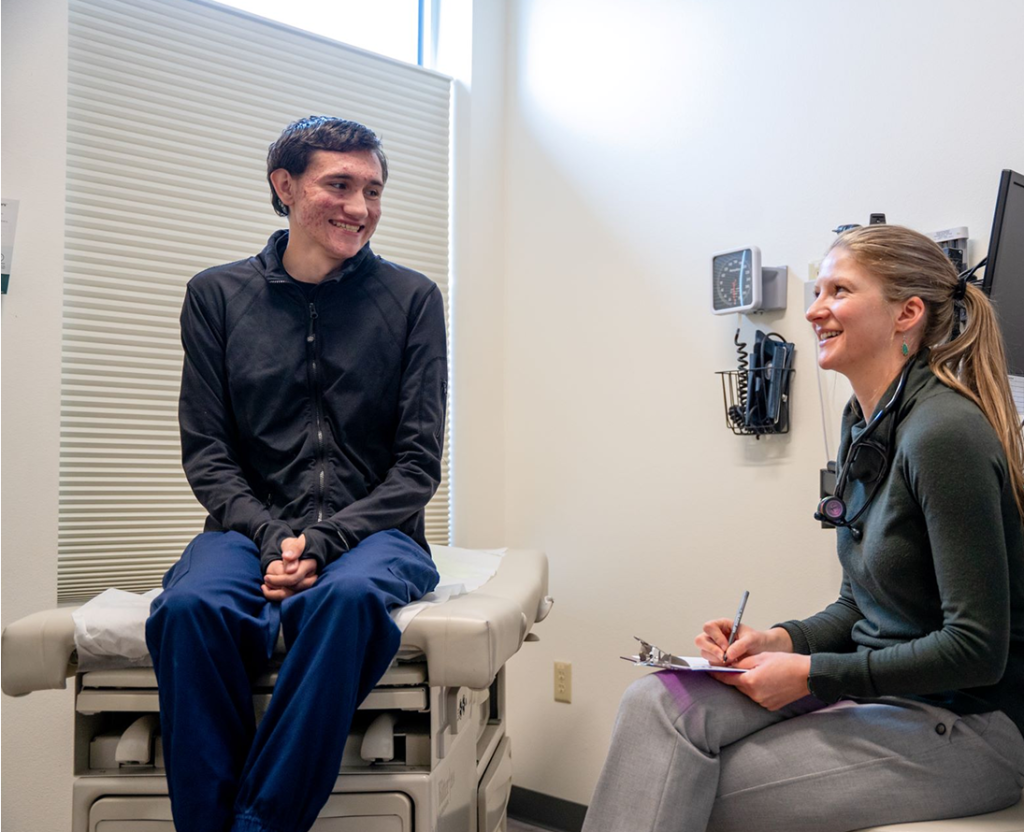 A young man sits on an exam table in a medical office, smiling at a healthcare professional who is taking notes on a clipboard. Medical equipment is visible on the wall behind them, reflecting the quality care found at Grand Junction Health Care.