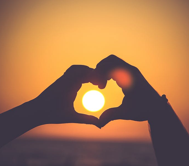 Two hands form the shape of a heart, framing the setting sun at sunset, representing the warmth and compassion found in Grand Junction Health Care against an orange sky and soft light in the background.