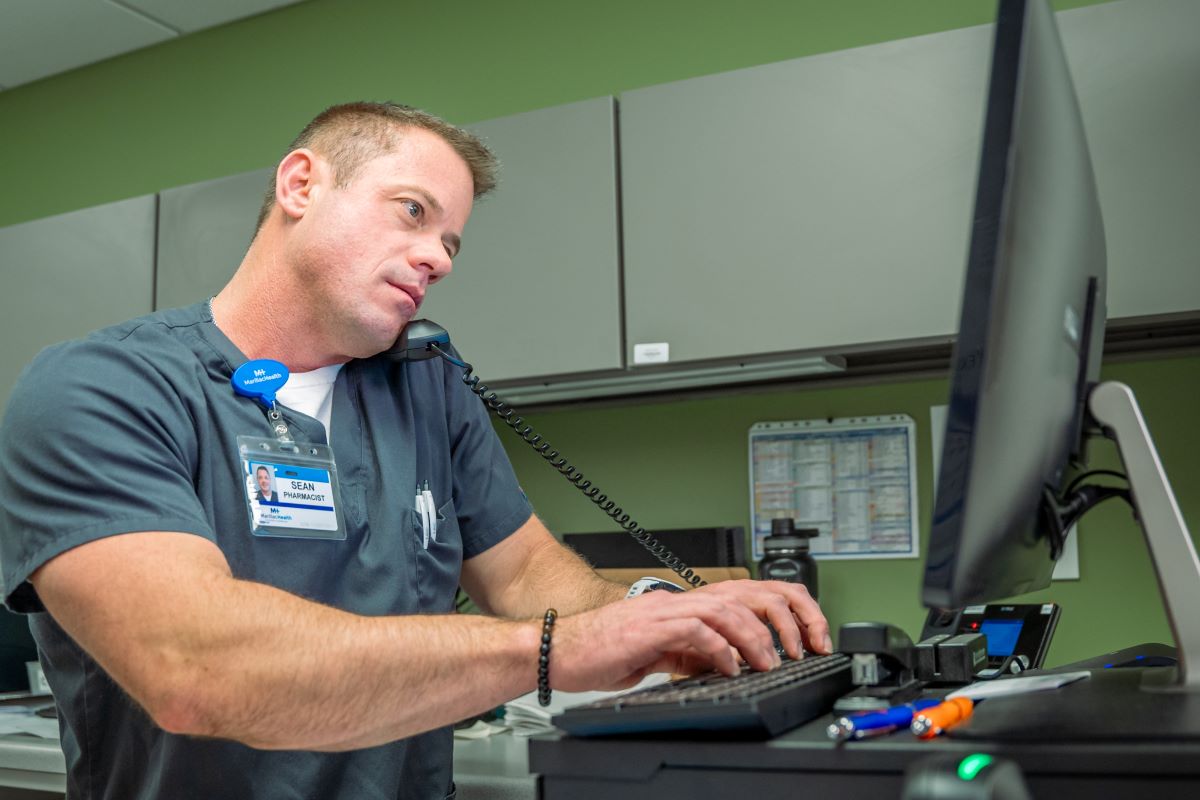 A Grand Junction Health Care worker multitasks at a desk, speaking on the phone while typing on a computer keyboard; pens and office supplies are scattered nearby, reflecting the busy pace of affordable health care.