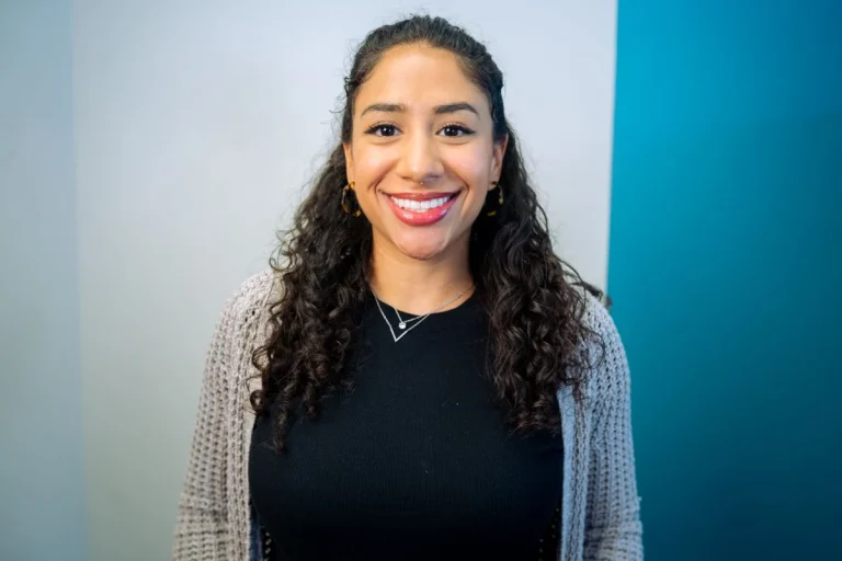 A young woman with curly dark hair, wearing a black top and gray cardigan, smiles at the camera. She stands in front of white and teal-colored walls at Montrose Health Care.