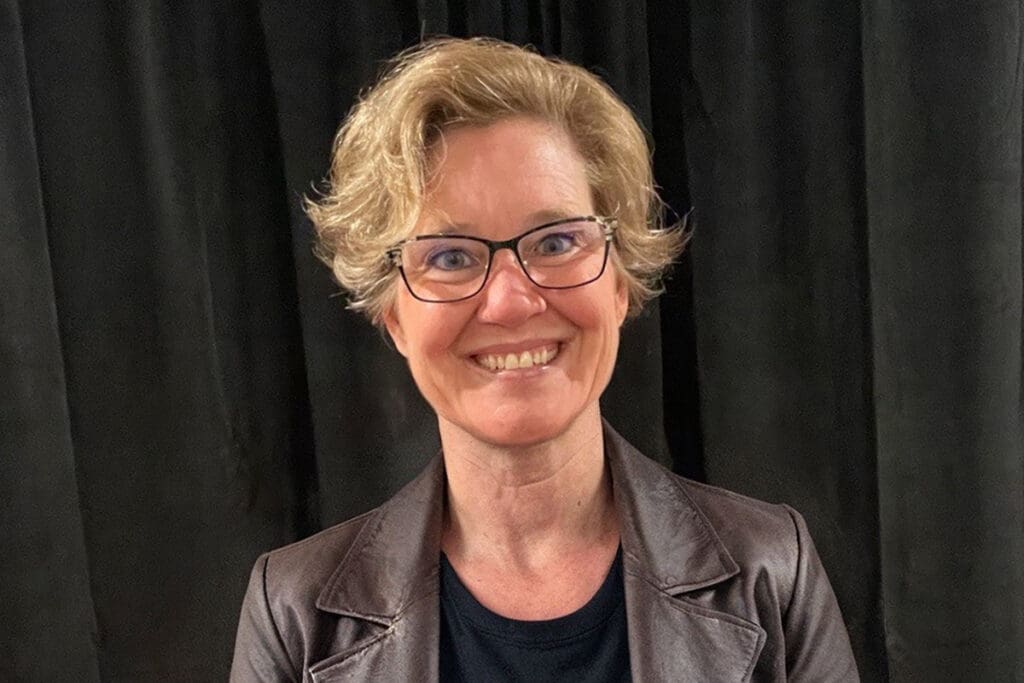 A middle-aged woman with blonde hair smiles at the camera. She is wearing a maroon V-neck shirt and stands in front of a background that is half white and half teal, reflecting confidence in affordable health care.