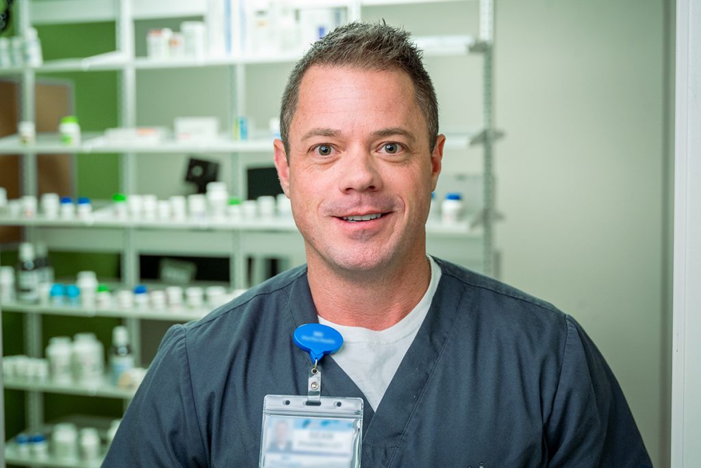 A man in medical scrubs with a name badge smiles in front of shelves filled with medication bottles, reflecting the welcoming atmosphere of Grand Junction Health Care in a pharmacy or medical setting.