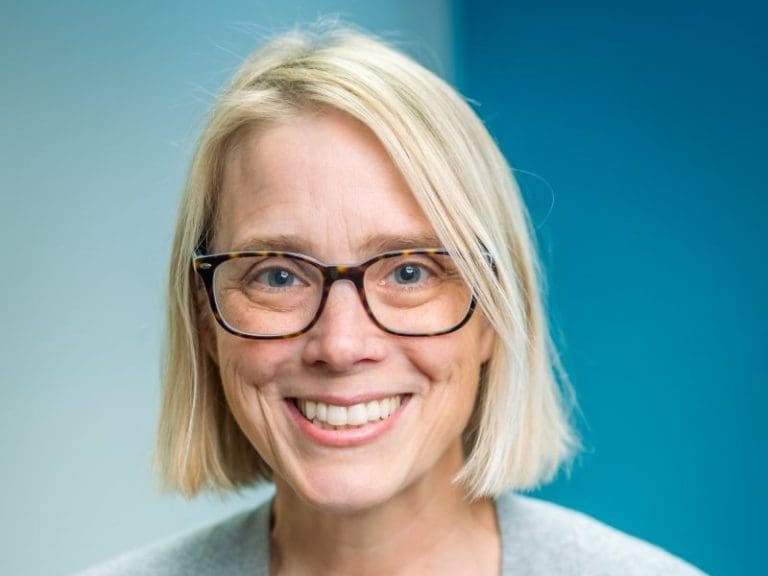 A smiling woman with straight blonde hair, wearing glasses and a grey top, stands in front of a blue and light blue background, representing Montrose Health Care.