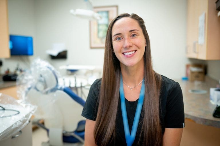 A woman with long brown hair wearing a black top and blue lanyard smiles in a Montrose Health Care dental office, with a dentist chair and medical equipment in the background.