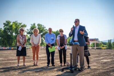 Six adults stand outdoors on a dirt lot, with one man in a suit speaking into a microphone at a podium about Affordable Health Care. The others listen attentively. Trees and a clear blue sky are in the background.