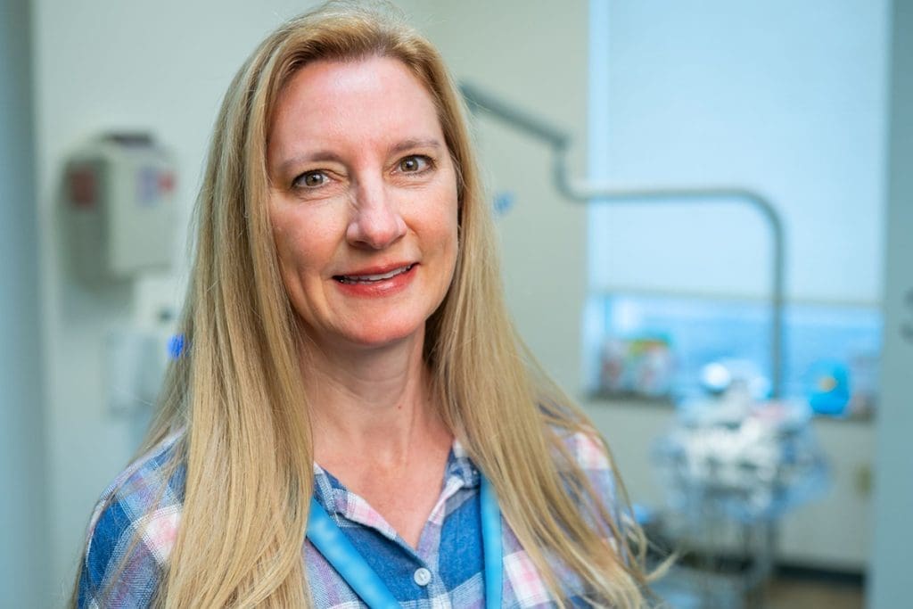 A woman with long blonde hair and a plaid shirt smiles in a bright Montrose Health Care dental office, with dental equipment and a window visible in the background.